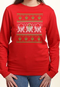Czerwony sweter z pikselowym wzorem droidów Star Wars R2-D2 oraz dwóch jednostek R2 w zielono-białym świątecznym wzorze, noszony przez osobę z rękami na biodrach.