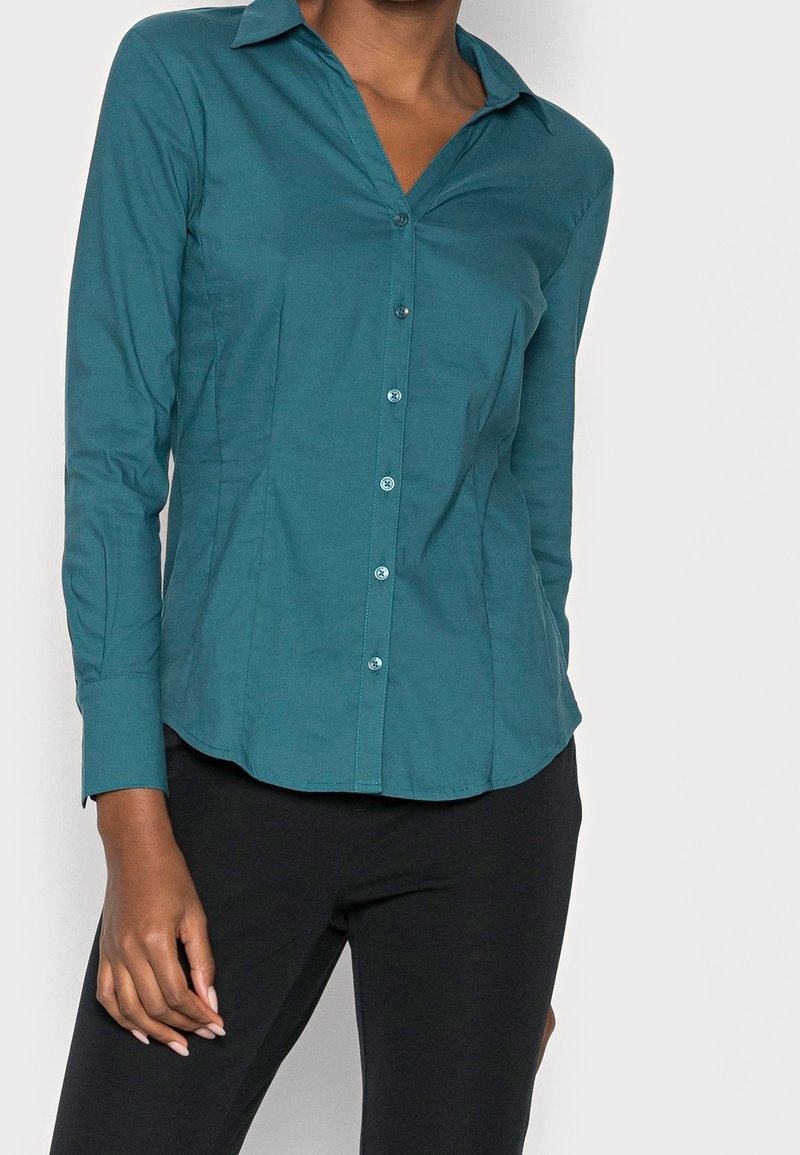 Camicia azzurra con bottoni realizzata in tessuto liscio, con maniche lunghe, colletto a punta e vestibilità sagomata, abbinata a pantaloni neri.