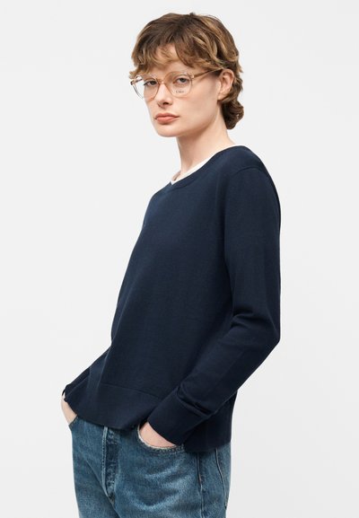 Personne aux cheveux courts et ondulés, portant des lunettes rondes transparentes, un pull bleu marine et un jean bleu, debout les mains dans les poches.