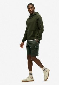 Oliwkowy hoodie z zamkiem błyskawicznym, w połączeniu z pasującymi szortami cargo. Beżowe sneakersy i skarpetki w niebieski wzór dopełniają całość.