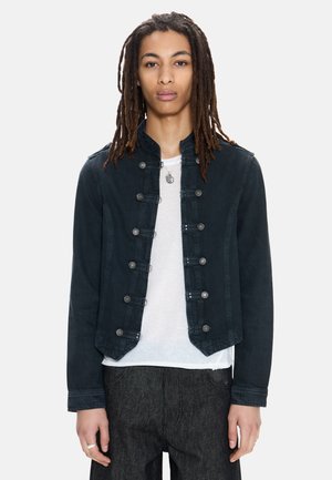 Mlada oseba z dolgimi rasta lasmi, oblečena v temno vojaško jakno, belo srajco, temne hlače in okrogel obesek na vrvici.