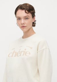 Krem sweatshirt med oversized passform, med stor "chérie" trykt i lys beige. Myk tekstur og rund hals. Subtile grafiske detaljer.