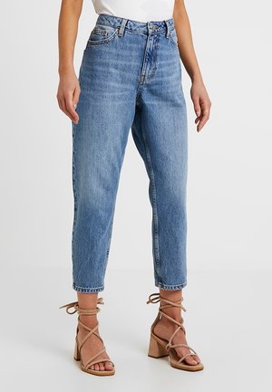 Femme portant un jean court taille haute bleu clair et des sandales beiges à talons blocs avec brides, debout sur un fond neutre.