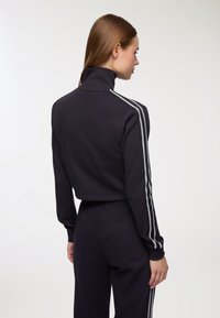 Pullover nero accorciato con colletto alto, caratterizzato da strisce bianche lungo le maniche. Texture liscia, design aderente, polsini elasticizzati.