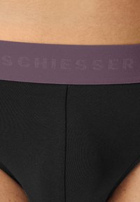 Zwarte onderbroek met een gladde textuur en een brede paarse elastische tailleband met veroutste merkletters.