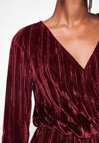 Vinfärgad sammetklänning med djup V-ringning, långa ärmar och mjuk, vertikal ribbad textur. Har en samlad midjedetalj.