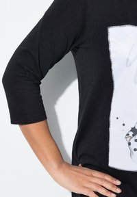 Schwarzes, T-Shirt mit drei Viertelärmeln und einem weißen Grafikdesign, das aus Punkten und einem genähten Umriss besteht. Glatte Textur, lockere Passform.