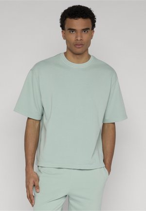 Mężczyzna o kręconych włosach, ubrany w luźną, jasnozieloną koszulę z krótkim rękawem i pasujące spodnie, stojący na jednolitym tle.