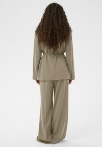 Gaiši olīvu zaļa uzvalka komplekts ar piegriezta stila žaketi ar jostu un plaša piegriezuma biksēm. Gluda tekstūra ar strukturētu dizainu un garām piedurknēm.