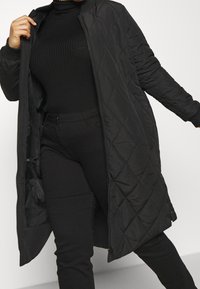 Person som bär en lång svart quiltad kappa, svart ribbad turtlenecktröja och svarta byxor, poserar med kappa öppen mot en vit bakgrund.