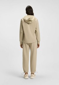 Completo activewear beige composto da una giacca con cappuccio e pantaloni a sigaretta. Tessuto liscio, polsini elasticizzati e corde regolabili alla caviglia. Design leggero e casual.