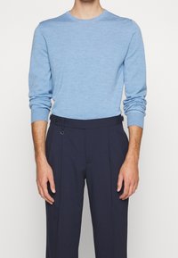 Homme portant un pull bleu clair à manches longues et un pantalon plissé bleu marine foncé, debout devant un fond uni.