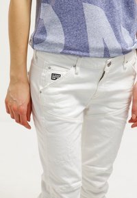 Pantalones de denim blanco con un ajuste slim, que cuentan con una pequeña etiqueta negra y un sutil detalle desgastado en la tela. Incluye acentos de herrajes metálicos.