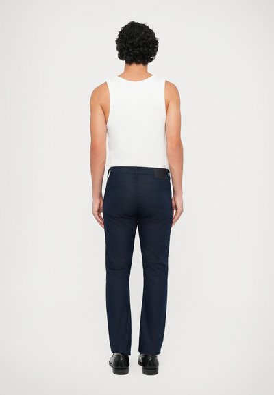 Calças de marinha com um corte slim, design de frente plana e uma textura suave, combinadas com uma camisa branca sem mangas; exibidas de costas.
