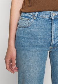 Los vaqueros de mezclilla azul claro presentan un diseño clásico de cinco bolsillos, costuras visibles y cierre de botón. La tela tiene una textura suave.