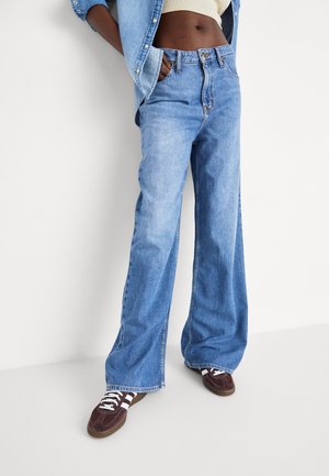 Persona que lleva vaqueros azules sueltos, un top corto de color claro, chaqueta vaquera y zapatillas marrones con rayas blancas, apoyada contra una pared blanca.