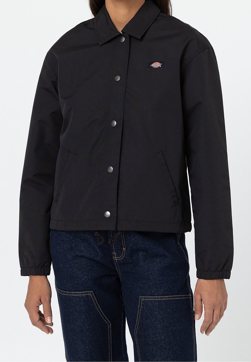 Personne portant une veste noire boutonnée avec col et petit logo sur la poitrine, associée à un pantalon en denim bleu foncé avec coutures visibles.