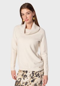 Pull beige avec un col cheminée, des poignets et un ourlet côtelés. Texture douce. Associé à une jupe à motifs dans les tons de marron et crème.