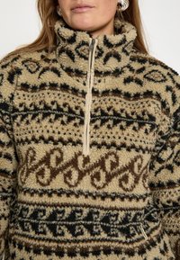 Bežový fleecový pulóver s hnedými a čiernymi vzorovanými detailmi. Obsahuje polovičný zips a vysoký golier pre dodatočné teplo.