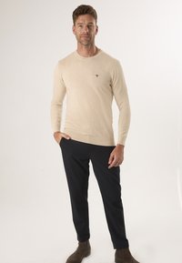 Beige gebreide trui met lange mouwen, ronde halslijn en subtiele branding op de borst, gecombineerd met donkere broeken en bruine schoenen.