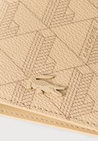 Επιφάνεια από μπεζ δέρμα με υφή, με εντυπωμένο γεωμετρικό σχέδιο και ένα μικρό ανάγλυφο έμβλημα κροκοδείλου κοντά στον ραμμένο ακμή.