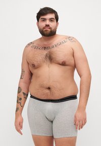 Boxer in grigio con una cintura nera che riporta la scritta "Jack & Jones". Il modello ha un tono di pelle chiaro e tatuaggi visibili sulle braccia.