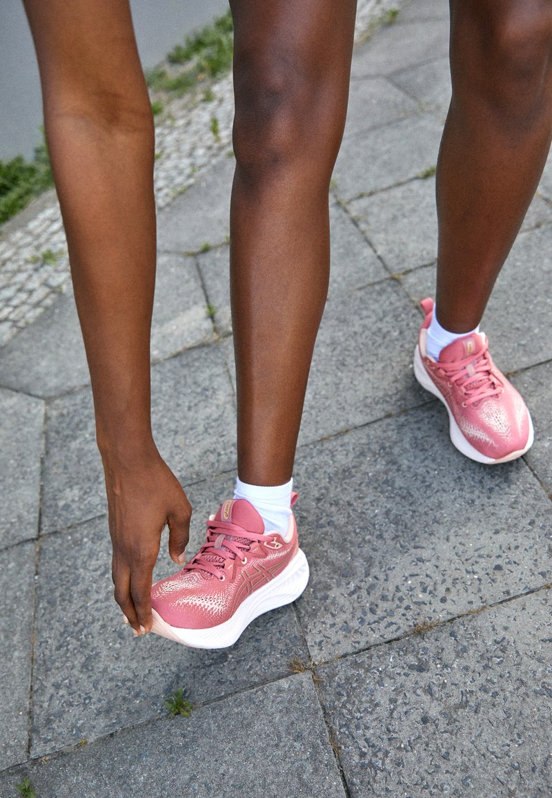 Rosa Sportschuhe mit einer strukturierten Oberseite, weißen Sohlen und Schnürsenkeln. Getragen mit weißen Knöchelsocken vor einem Hintergrund aus Steinpflaster.