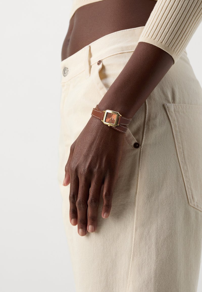 Montre à cadran carré en or avec un bracelet en cuir marron et un cadran aux accents orange, portée sur une main reposant contre un tissu de couleur crème.