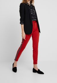 Zwarte blazer, gestreepte zwarte blouse, rode broek met zijstrepen, en zwarte loafers. Slank model met ruches aan de kraag.