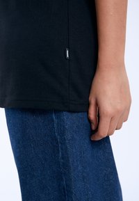 Camiseta de algodón negra con etiqueta en la costura lateral, combinada con jeans de mezclilla azul. La camiseta tiene un diseño simple y una textura suave.
