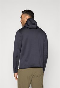 Granatowy sweatshirt z kapturem o gładkiej fakturze, dopasowanym kroju i kontrastowych szwach, w zestawieniu z khaki spodniami. Wyświetlony widok z tyłu.