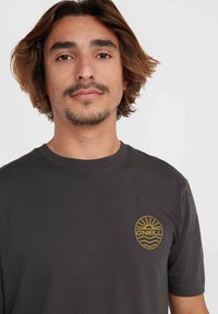 Camiseta gris oscuro de algodón. Presenta un logo circular en color beige con un diseño de sol y ola ubicado en la parte izquierda del pecho. Mangas cortas.