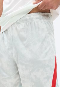 Ανδρικό χέρι σηκώνει λευκό πουκάμισο αποκαλύπτοντας ανοιχτό γκρι αθλητικό σορτς με διακριτικό σχέδιο και κόκκινη ρίγα στην πλαϊνή τσέπη.