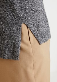 Grauer Strickpullover mit seitlichem Schlitz, kombiniert mit hellbraunen Stoffhosen. Der Pullover weist ein strukturiertes Muster und einen gerippten Saum auf.