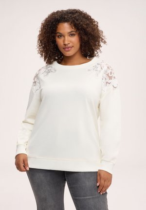 Kvinde med krøllet hår iført en hvid langærmet top med broderede transparente skulderpaneler og grå jeans, stående mod en lys baggrund.