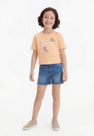 Κορίτσι που χαμογελάει με κοντά σκούρα μαλλιά, φοράει ένα ροδακινί μπλουζάκι με λουλουδάτο σχέδιο, τζιν σορτς και μπεζ παπούτσια slip-on, που στέκεται μπροστά σε λευκό φόντο.