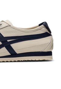 Beżowe skórzane sneakersy z granatowymi akcentami i paskami, charakteryzujące się teksturowaną powierzchnią, kontrastującą podeszwą oraz niskoprofilowym designem.