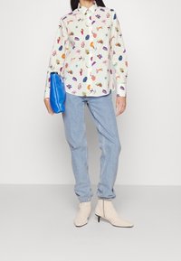 Camisa blanca de manga larga con patrones abstractos multicolores, combinada con vaqueros azul claro y botines beige texturizados. Bolso de mano azul.