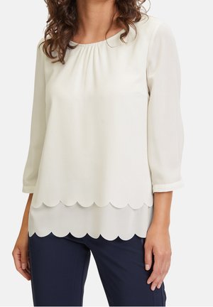 Vrouw draagt een witte blouse met een geschulpte zoom en driekwart mouwen, gecombineerd met donkerblauwe broek, tegen een effen achtergrond.