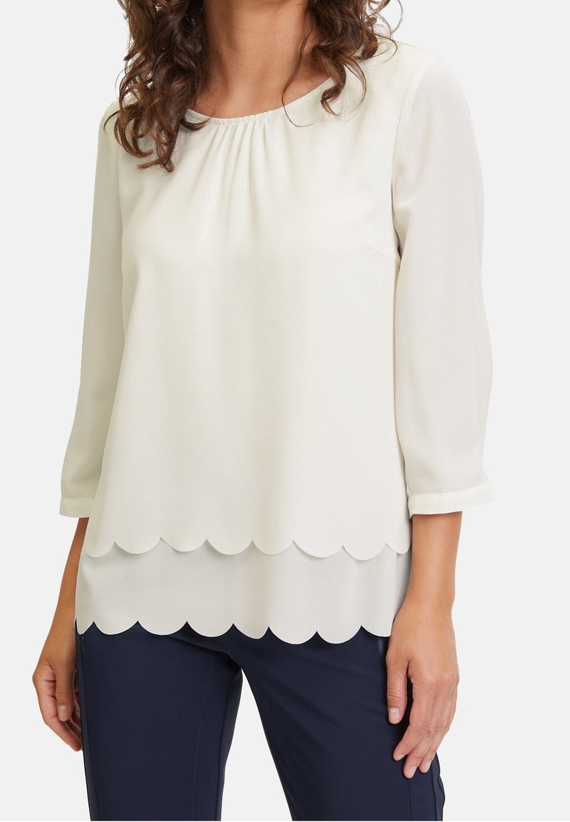 Femme portant un chemisier blanc à ourlet festonné et manches trois-quarts, associé à un pantalon bleu foncé, sur un fond uni.