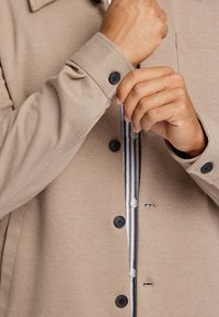 Beige button-down Hemdjacke aus strukturiertem Stoff mit schwarzen Knöpfen und einem gestreiften Hemd darunter. Sichtbare verstellbare Manschettendetails.