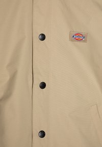 Μπεζ ζακέτα με λείο ύφασμα, διαθέτει μαύρα κουμπιά με σ snap και πολύχρωμο λογότυπο "Dickies" ραμμένο στο αριστερό στήθος.