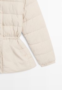 Giacca beige imbottita con vita stretta, polsini elasticizzati e motivo a pois rettangolari. Texture morbida con tasche laterali.