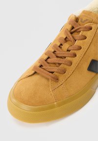 Sneakers de suedă maro cu o suprafață texturată, șireturi maro și un accent logo negru. Prezintă o talpă din cauciuc rotunjită, cu un pattern pentru aderență.