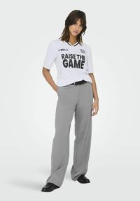 Biała koszula z krótkim rękawem z napisem "RAISE THE GAME" wydrukowanym na czarno, w połączeniu z jasnoszarymi, szerokimi spodniami oraz czarnymi butami.