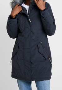 Personne portant un parka bleu marine foncé avec une capuche bordée de fourrure, fermeture éclair à l'avant, deux poches latérales zippées et un jean bleu clair.