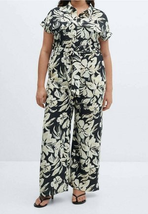 Femme portant une combinaison noire à imprimé floral blanc, manches courtes, boutons à l'avant, ceinture à la taille, pantalons larges, et sandales à talons noires.