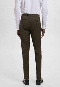 Man in een donkerbruin slim-fit broek, wit langarmshirt en zwarte schoenen, staand met zijn rug naar de camera tegen een effen achtergrond.