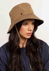 Vrouw met lang, donker haar, die een tan emmerhoed met een kinband en een marineblauwe jas draagt, kijkend naar de zijkant tegen een effen achtergrond.