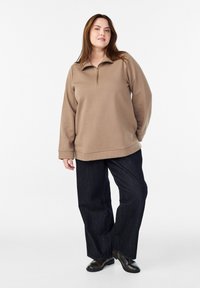 Brązowa bluza zapinana na zamek, wykonana z miękkiego materiału, z kołnierzem stójkowym. Połączona z ciemnoniebieskimi spodniami dzwonami i czarnymi botkami za kostkę.